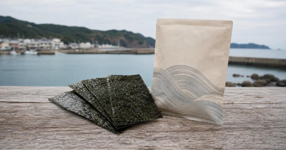 静かな海辺を背景に無地の海苔袋と板海苔を置き、海の町の印象と商品の原料産地が別であることを表したイメージ